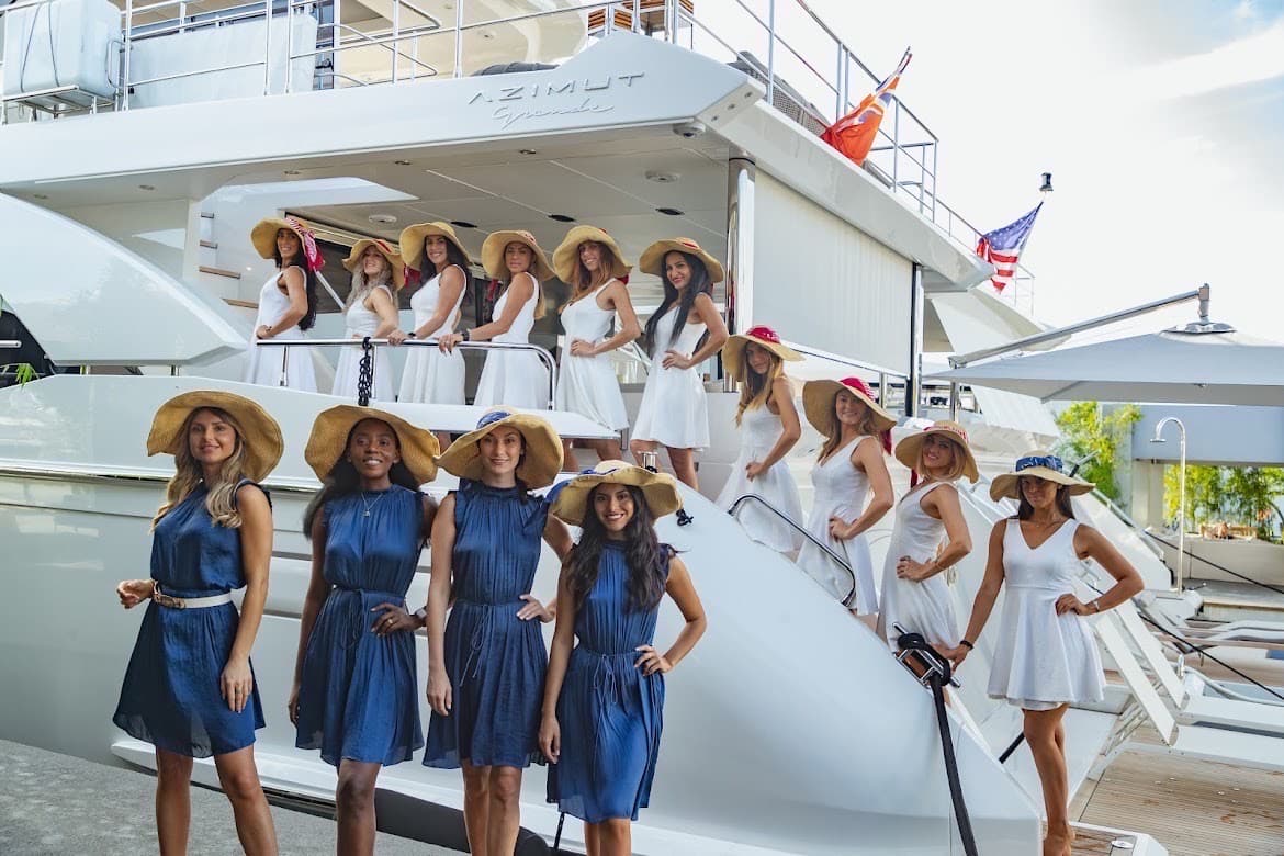 More than a dozen models in white and navy dresses with wide-brim sun hats pose aboard and beside an Azimut Yachts vessel at a dockside event, the AZIMUT wordmark visible on the hull.