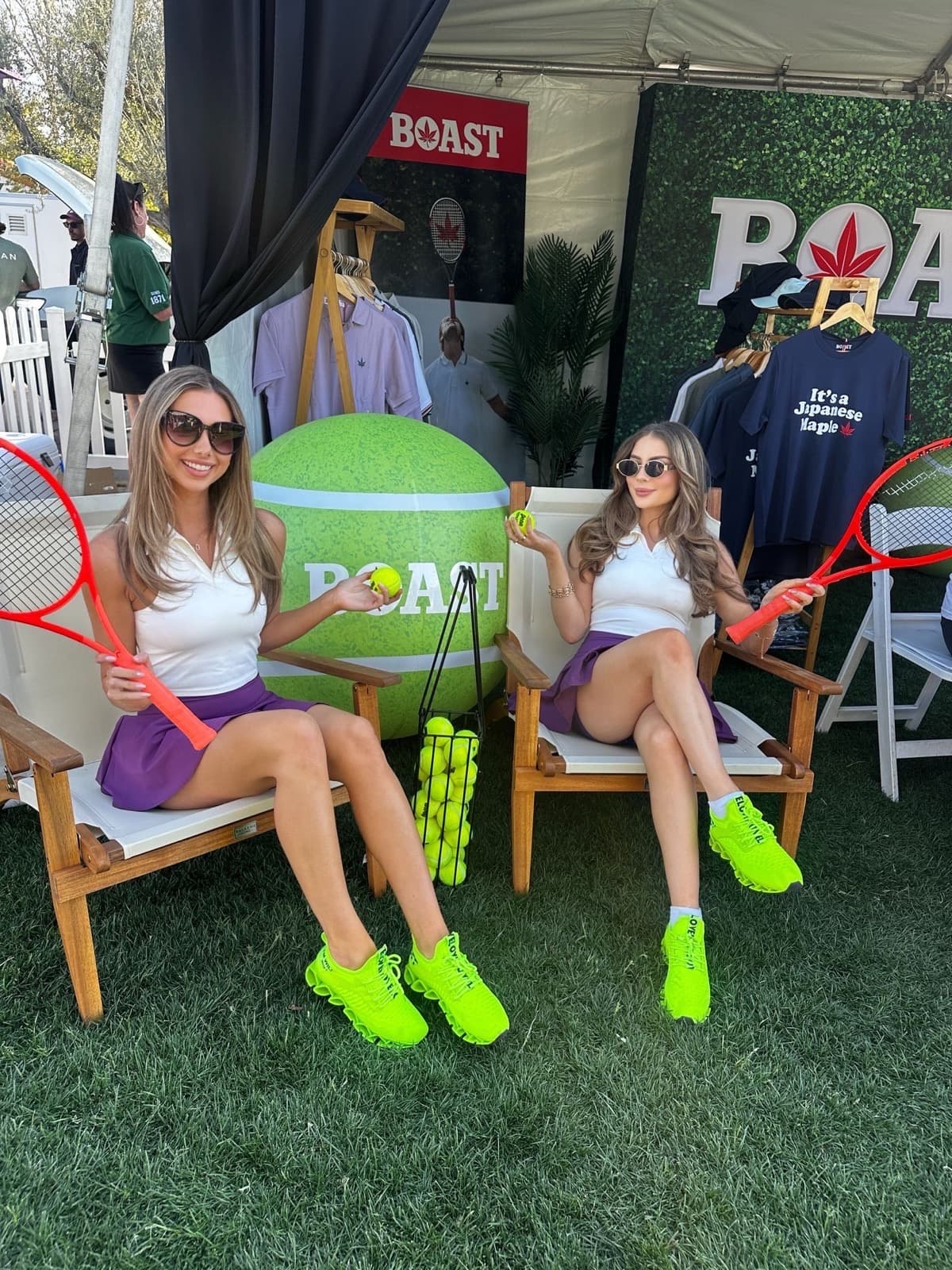 Two brand ambassadors in white polos and purple skirts pose with tennis rackets at a BOAST apparel trade show booth.