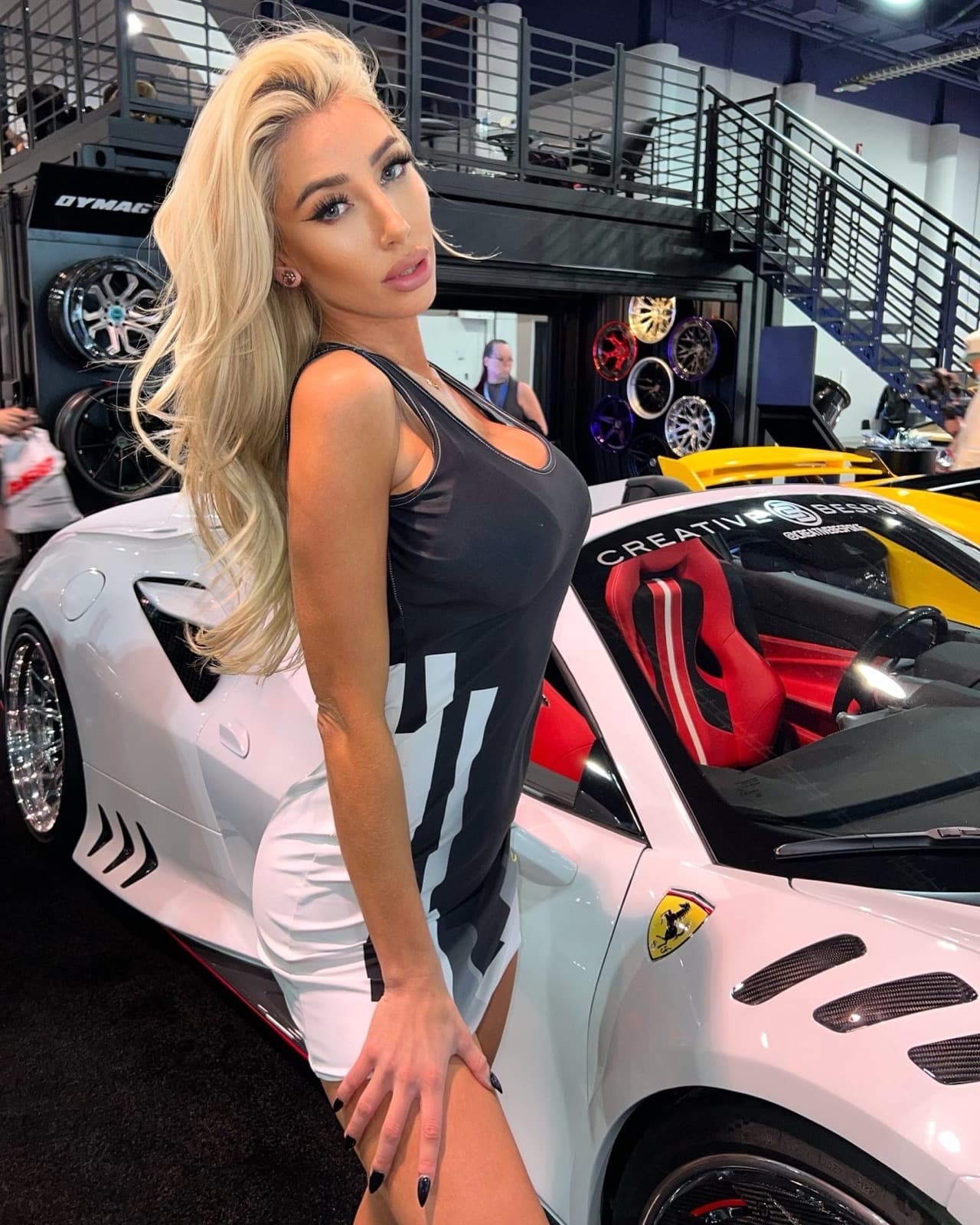 A brand ambassador in a black and white tank dress poses beside a white Ferrari at a Creative Bespoke car show exhibition booth.