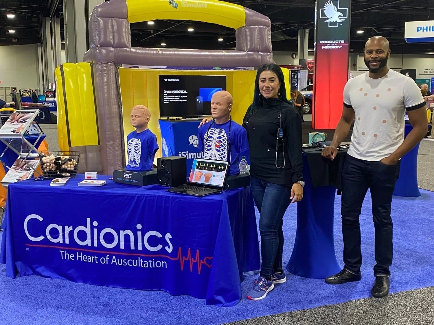 A brand ambassador in a black turtleneck and a colleague in a printed anatomy t-shirt staff the Cardionics booth, its table banner reading 'Cardionics — The Heart of Auscultation,' beside pediatric and adult auscultation-training mannequins at a medical trade show.