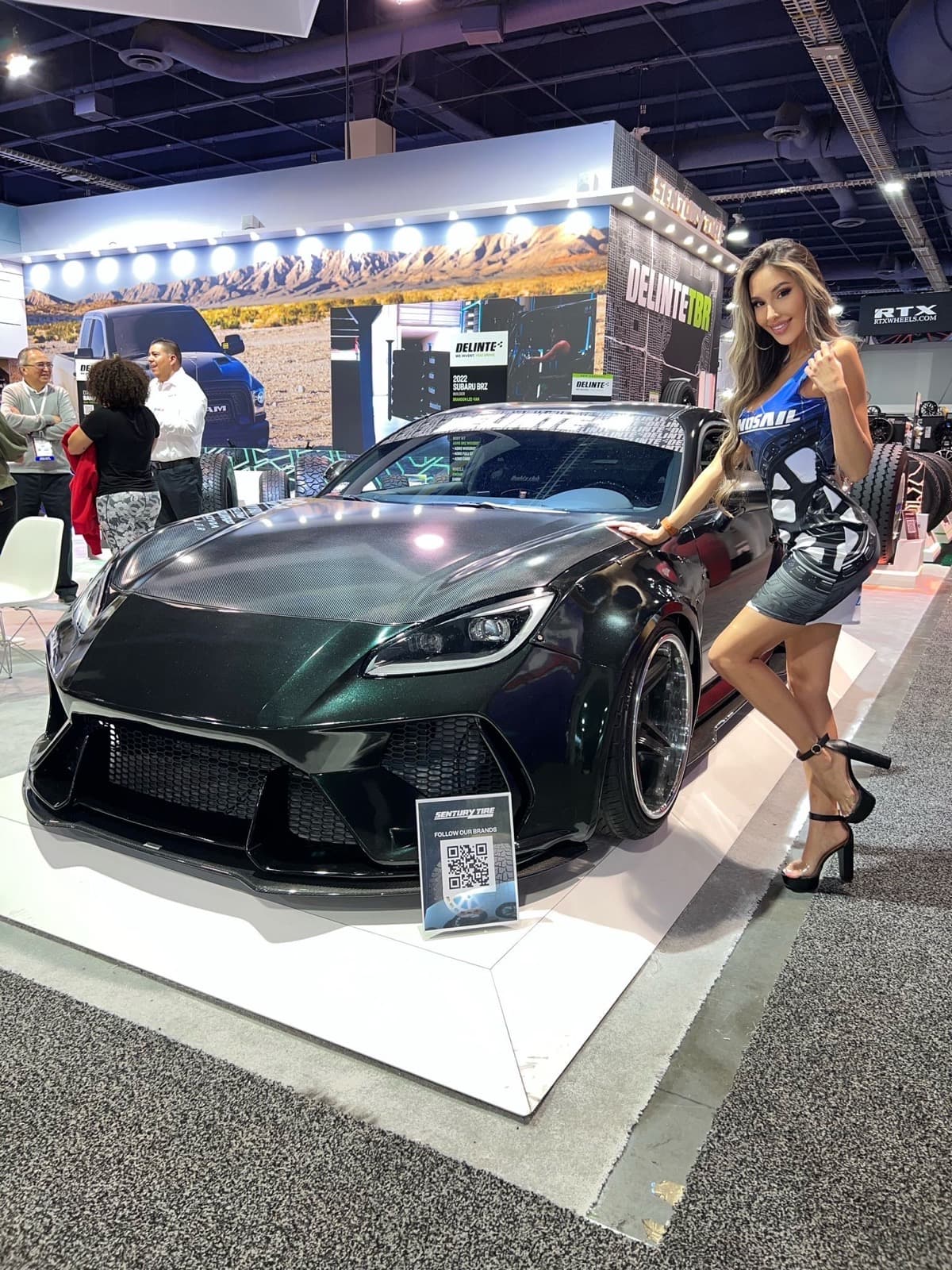 A brand ambassador poses beside a dark green sports car at a Delinte/Sentury Tire trade show booth.
