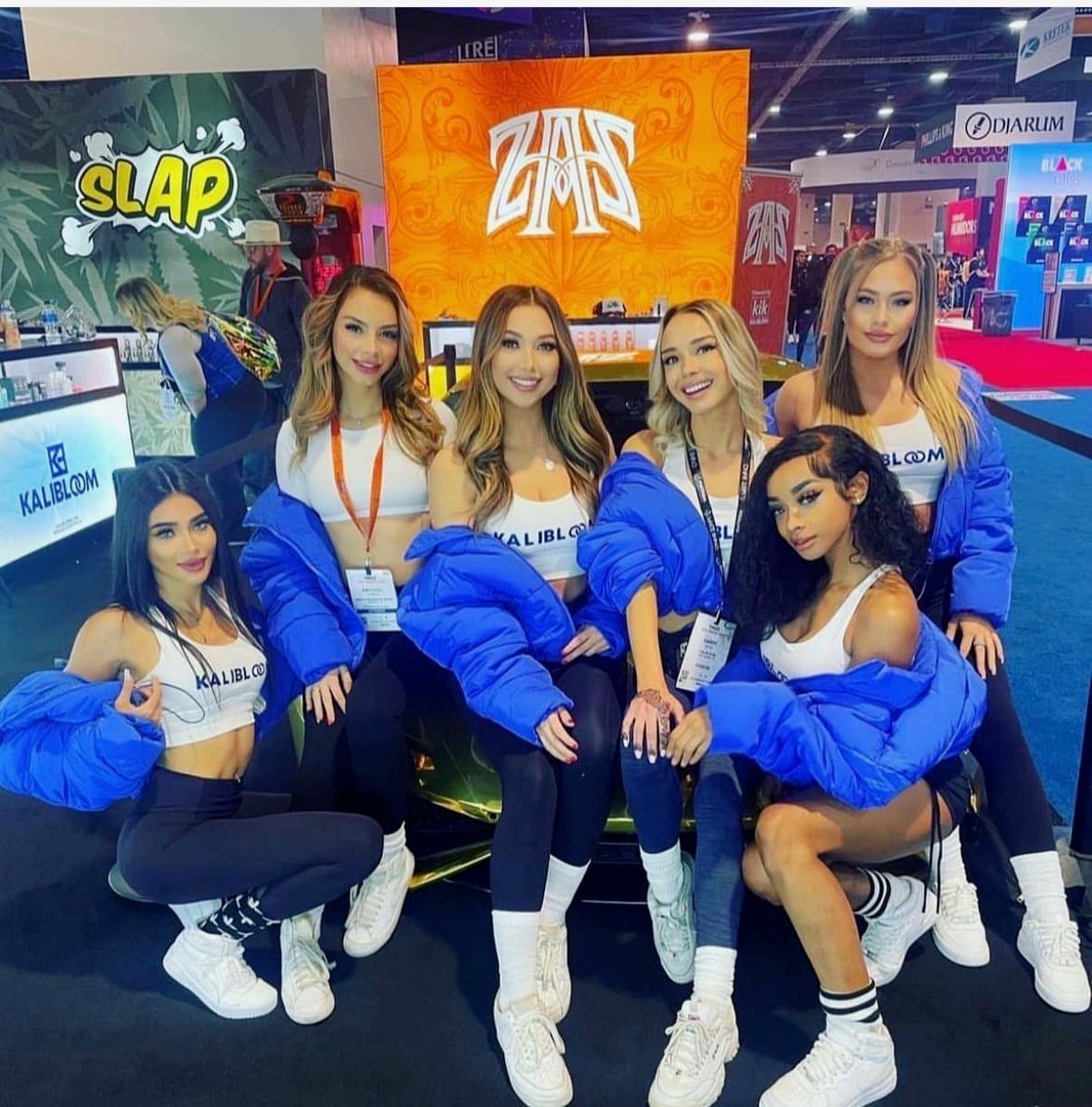 Six brand ambassadors in white Kalibloom crop tops and blue puffer jackets pose beside a branded car at a trade show booth backed by SLAP and Kalibloom signage.