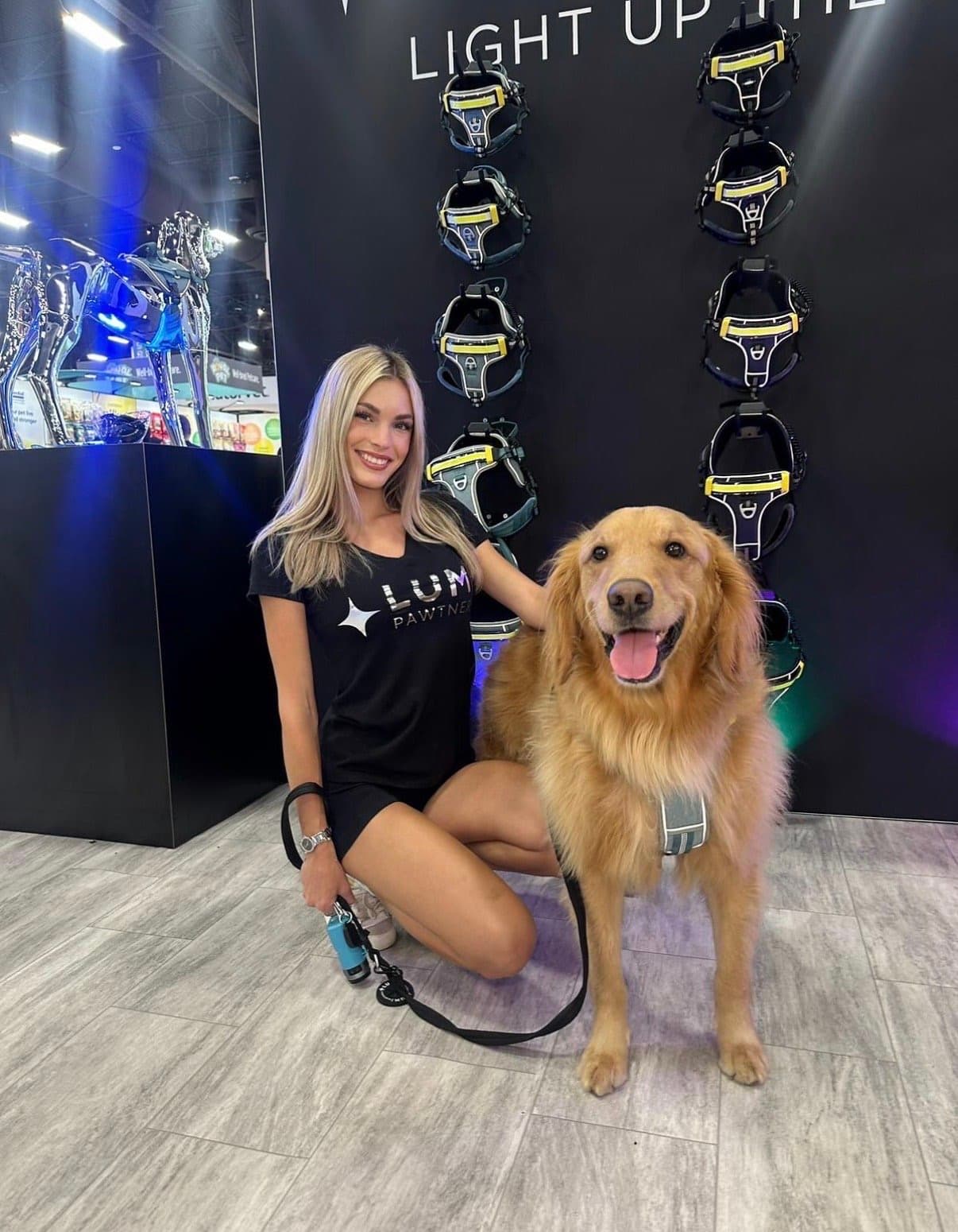 A brand ambassador poses with a golden retriever in front of light-up pet harnesses at a LUM Pawtner trade show booth.
