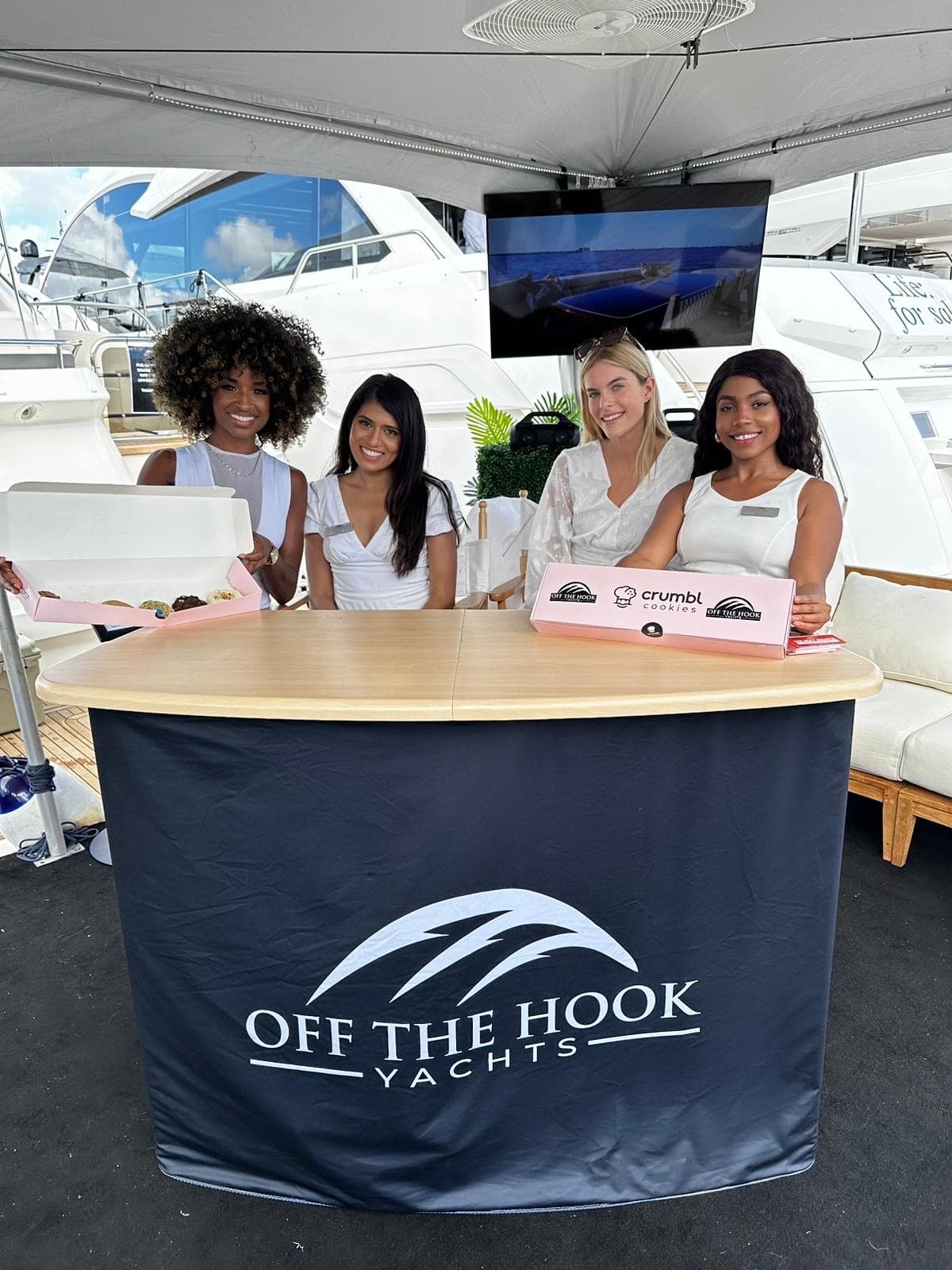 Four brand ambassadors in white outfits staff an Off The Hook Yachts booth with Crumbl Cookies boxes on the table.