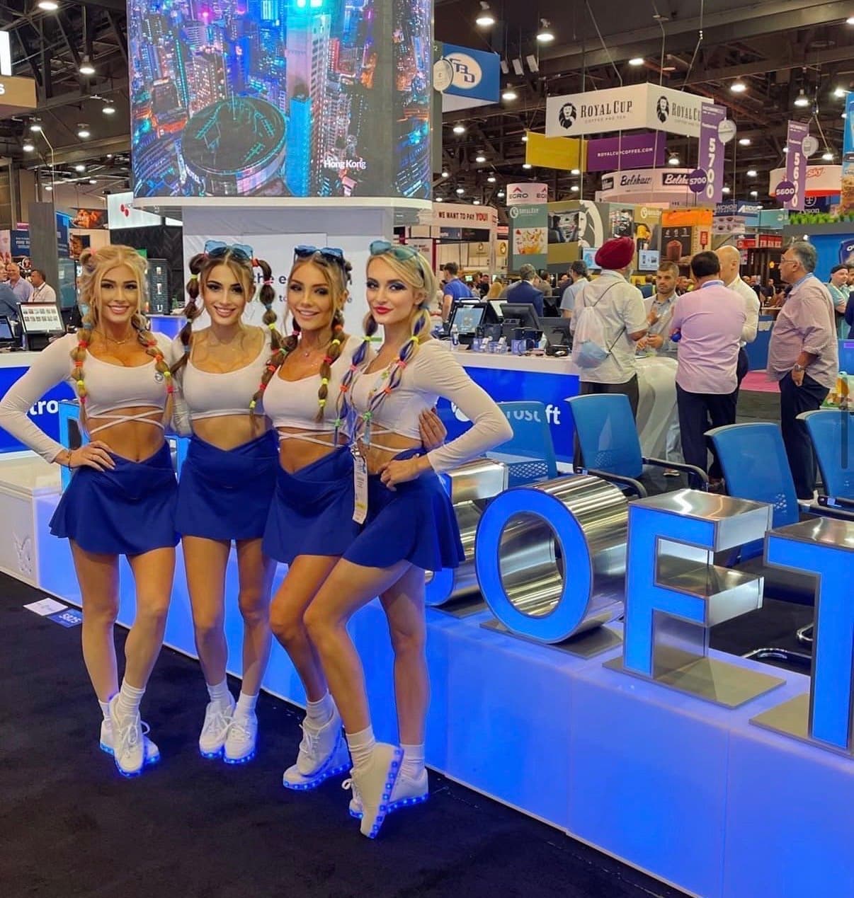 Four brand ambassadors in white wrap crop tops, navy skirts, and colorful braided pigtails pose at a Petrosoft trade show booth, the counter behind them reading "...rosoft" beside large illuminated letters spelling "...OFT" and a screen showing an aerial night view of Hong Kong, with Royal Cup Coffee signage visible on the show floor.