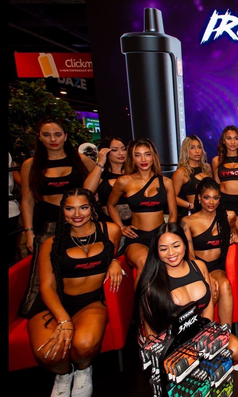 Seven brand ambassadors in matching black 'Iron Mike' crop tops pose together holding product 3-packs at the Iron Mike Tyson vape trade show booth, with Mike Tyson and his backdrop signage cropped out of frame.